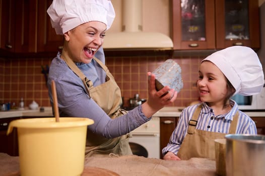 Cheerful mother and cute daughter have fun together, enjoying festive atmosphere in kitchen while cooking Easter cake