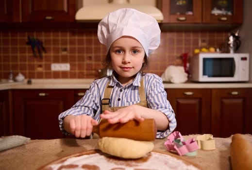 Authentic portrait of a lovely little child girl using rolling pin, rolls out dough on wooden board, smiling at camera
