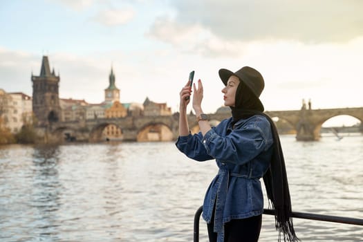 Stylish young woman traveller wearing black hat sitting taking pictures with her smartphone on Vltava river shore in Prague with Charles Bridge on background.