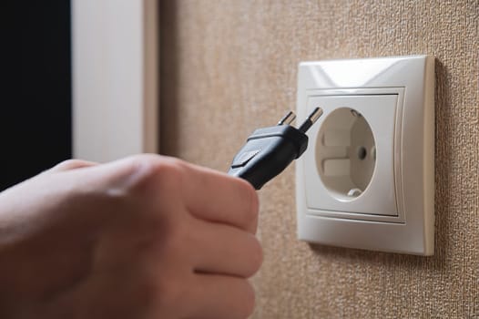 black cord with a plug from an electrical appliance is brought to the socket by hand, close-up