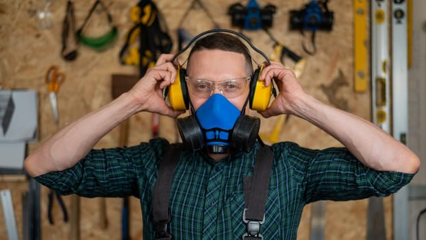 A carpenter in goggles and a respirator puts on noise-protection earmuffs.