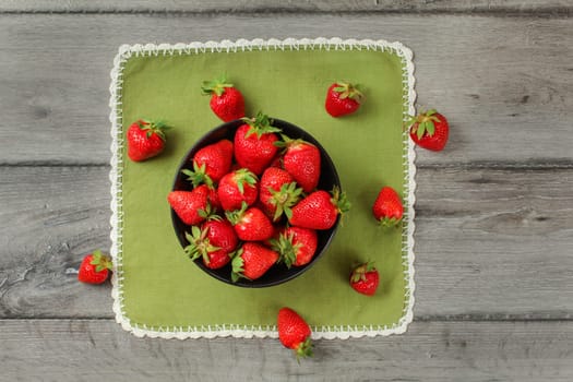 Tabletop view, small black ceramic bowl, with strawberries, some of them spilled on green tablecloth under.
