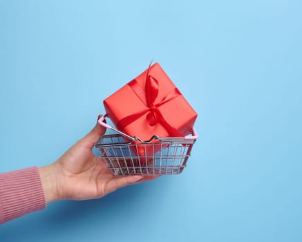 A woman's hand holds a red gift box wrapped in mid-air against a blue background, concept of celebration, surprise