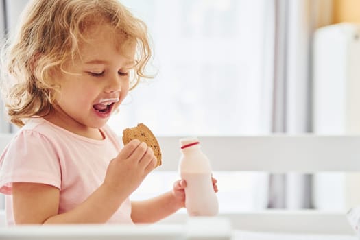 Sitting on the bed, eating cookies and drinking milk. Cute little girl in casual clothes is indoors at home at daytime