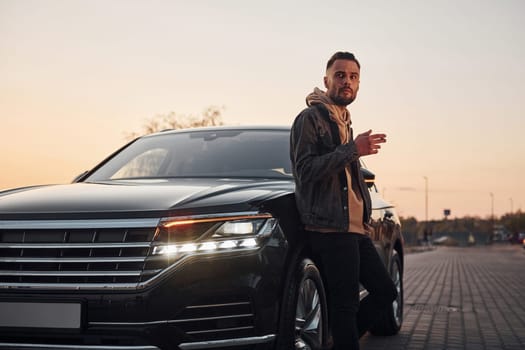 Handsome unshaved man in fashionable clothes standing near his black car and smoking