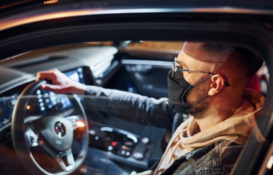In black protective mask. Handsome unshaved man in fashionable clothes riding his automobile