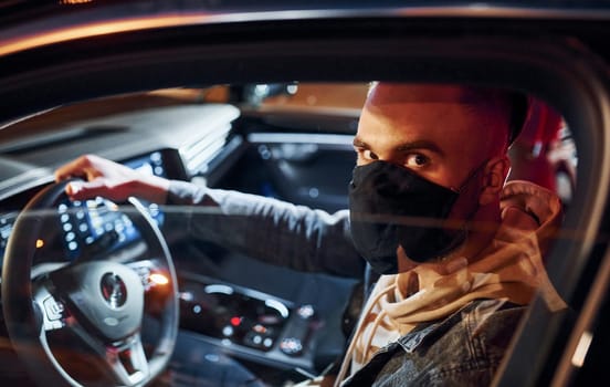 In black protective mask. Handsome unshaved man in fashionable clothes riding his automobile