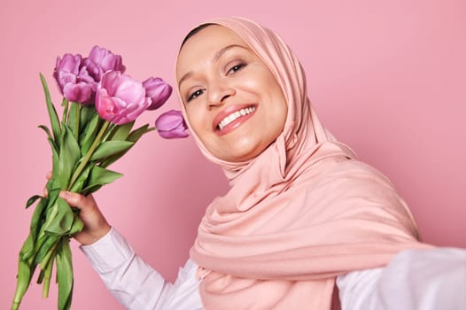 Self-portrait of beautiful positive happy smiling Middle-Eastern Muslim woman in pink hijab, holding bouquet of purple tulips for Mother's Day or Interntional Women's Day, isolated on pink background