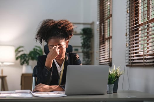 Young African American woman with afro hairstyle looks annoyed and stressed, sitting at the desk, using a laptop, thinking and looking at the camera, feeling tired and bored with depression problems
