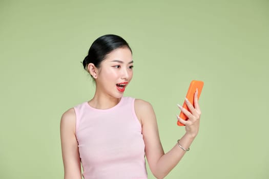 Young beautiful Asian woman feeling happy or surprise shock and holding smartphone over green background