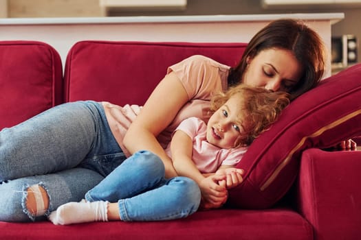 Lying down on red sofa. Young mother with her little daughter in casual clothes together indoors at home