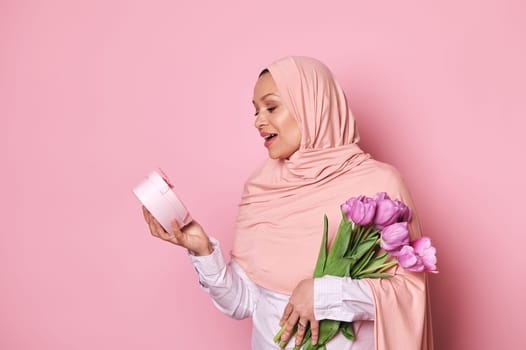 Muslim pretty woman in pink hijab, holds a bouquet of tulips, expresses amazement getting a presen for festive occasion,