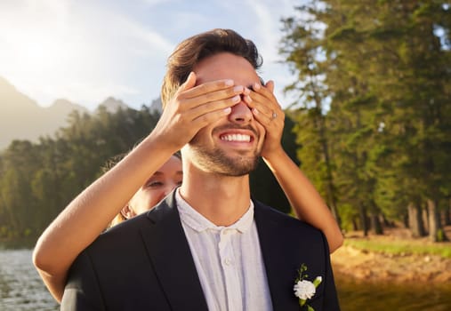 Love, wedding or surprise with a bride and groom by a lake, in celebration of a ceremony of tradition. Romance, marriage and hands over face with a married couple playing or joking together outdoor.