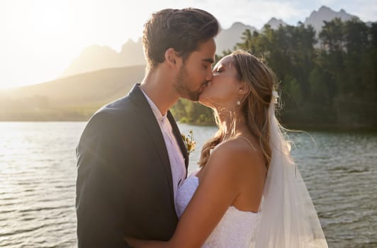 Love, wedding and a newlywed couple kissing by a lake outdoor in celebration of their marriage for romance. Water, summer or kiss with a bride and groom bonding together in tradition after ceremony.