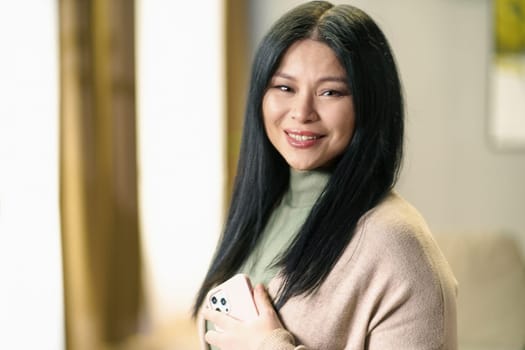 Mature and positive Asian woman with thick, luscious hair holds her smartphone in her hand, smiling with contentment and confidenced