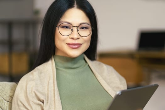 cheerful middle-aged Asian woman wearing glasses enjoys browsing internet on her tablet while sitting comfortably at home