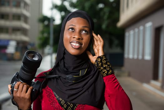 young muslim woman standing outdoors with a camera thinking about looking up there.