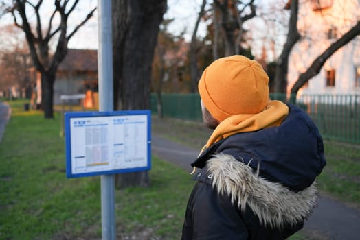 Guy in winter coat and yellow hat looking at a bus stop timetable schedule. High quality photo