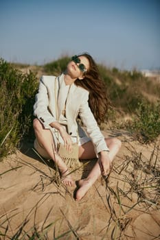 a beautiful woman in a light, fashionable jacket, in sunglasses sits barefoot on the sand in windy weather. High quality photo