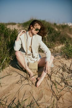 a beautiful woman in a light, fashionable jacket, in sunglasses sits barefoot on the sand in windy weather