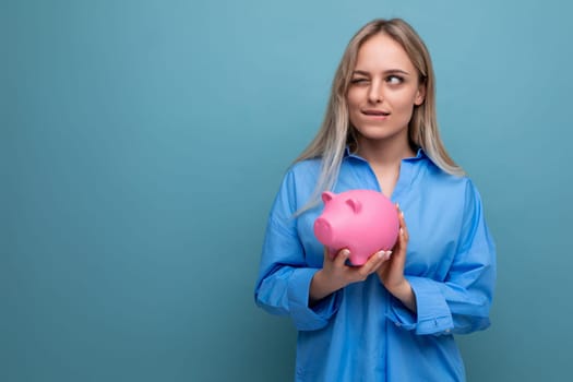 smart blond girl holding a piggy bank in the form of a pig with money on a blue isolated background