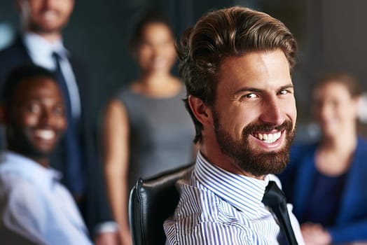 Success takes a team effort. Portrait of a smiling businessman sitting in an office with colleagues in the background.