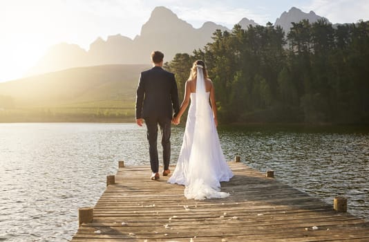 Love, wedding and back of couple by a lake standing, bonding and holding hands on the pier. Nature, romance and young bride and groom in an intimate moment together outdoor on a romantic marriage day