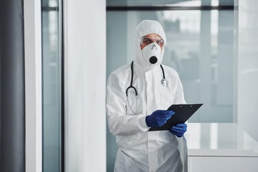 Male doctor scientist in lab coat, defensive eyewear and mask holding notepad in hands