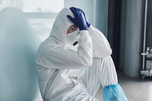 Tired doctor scientist in lab coat, defensive eyewear and mask sits and takes break