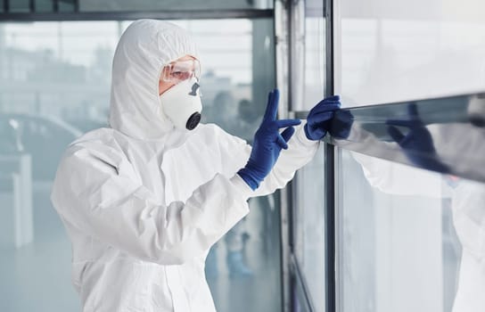 Male doctor scientist in lab coat, defensive eyewear and mask shows gesture through the window