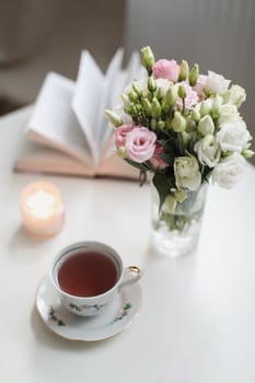 a spring still life, a bouquet of roses in a vase, a cup of tea and an book on white table. spring greeting card with spring flowers. Romantic background