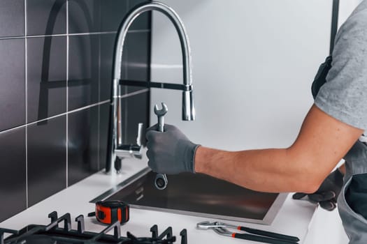 Close up view of young professional plumber in grey uniform holding wrench in hand on the kitchen