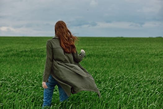a red-haired woman in a long raincoat stands in a green field in cloudy weather in a strong wind. High quality photo