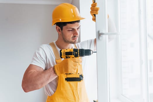 Handyman in yellow uniform installs new window by using automatic screwdriver. House renovation conception