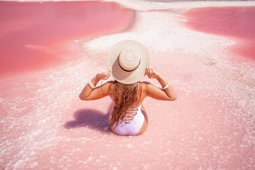 A woman traveler looks at an amazing pink salt lake. He sits with his back in a bathing suit and holds his hat in his hands. Wanderlust travel concept
