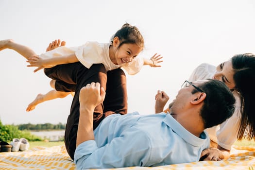 Family bonds over laughter and joy in the park. Father lifts his daughter high, as she happily flies like a plane with arms spread wide, and mother watches with pride. A picture of family fun and love