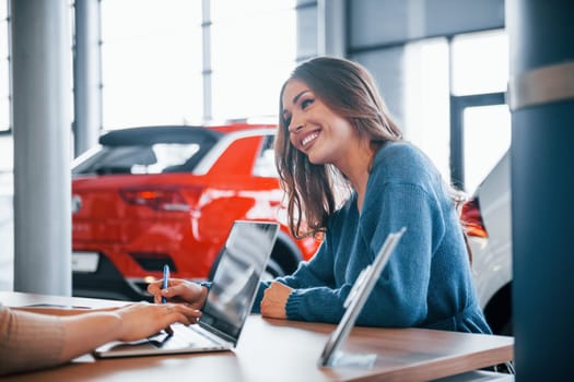 Cheerful woman sits with assistant in car shop and buying new vehicle