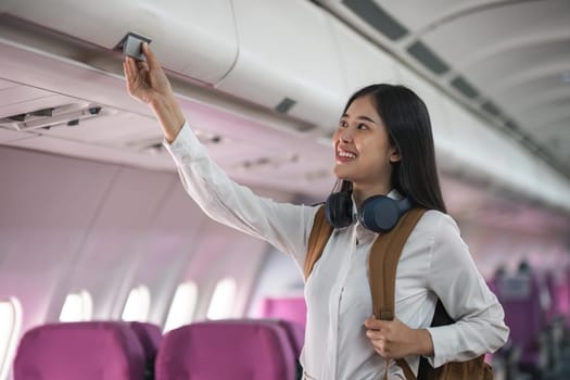 Young asian woman travel by airplane. Passenger putting hand baggage in lockers above seats of plane.