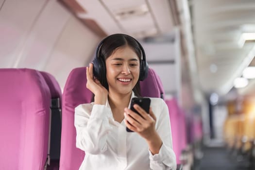 Cheerful female passenger in headphones for noise cancellation watching online movie during intercontinental flight in cabin of aircraft, happy young woman using wifi connection on board