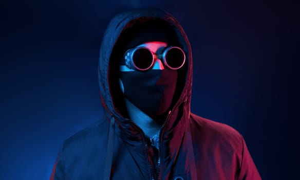 In mask, eyewear and hood. Neon lighting. Young european man is in the dark studio