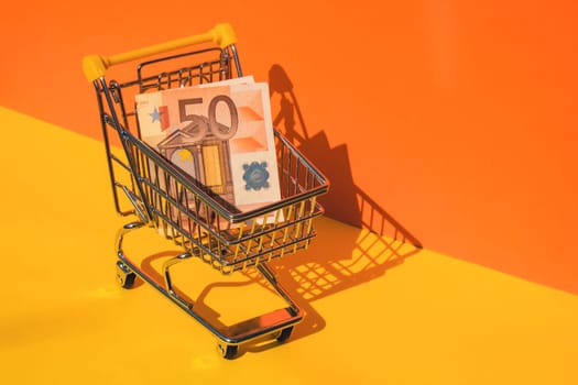 Euro paper currency 50 banknote money Shopping trolley cart on colorful orange yellow background. Copy space for your text. Online shopping, buy mall market shop consumer concept. Small toy supermarket grocery push cart. Food crisis. Rising food cost, grocery prices