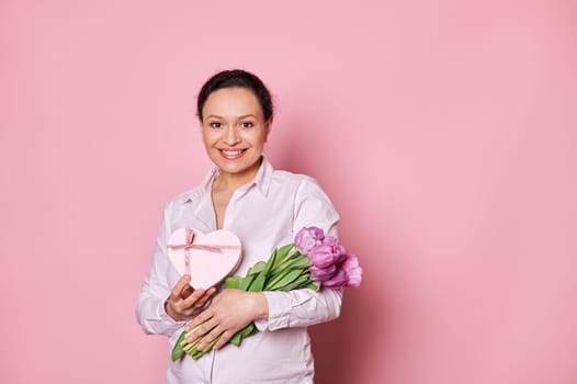 Authentic portrait on pink background of middle-aged smiling beautiful woman with gift box and bouquet of purple tulips