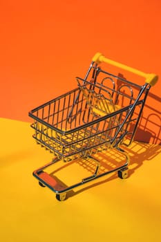 Empty shopping trolley cart on colorful orange yellow background. Copy space for your text. Online shopping, buy mall market shop consumer concept. Small toy supermarket grocery push cart. Food crisis. Rising food cost, grocery prices