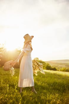 a woman in a long light dress walks through the countryside in a hat and with a basket in her hands in the rays of the setting sun enjoying nature