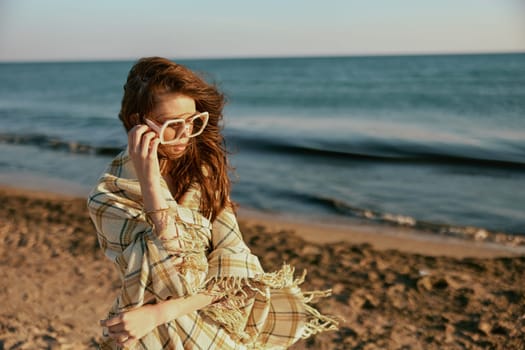 a woman wrapped in a plaid walking along the sea coast straightens her sunglasses