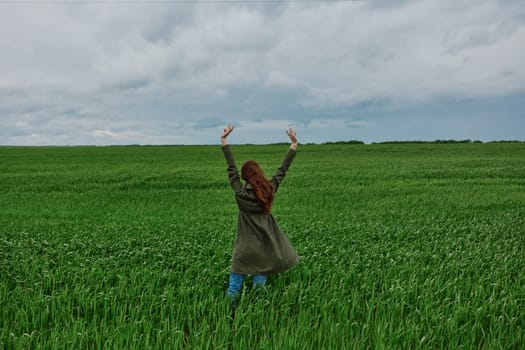 a woman in a long coat stands in a green field on a cloudy day with her hands up to the sky