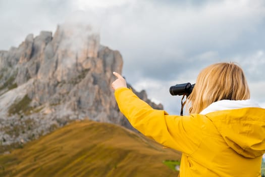 Blonde woman looks at Passo Giau pass through binoculars pointing finger on rocky mountain covered with clouds. Female tourist enjoys hiking in Italian Alps