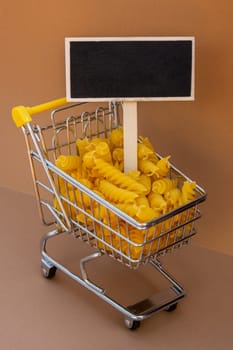 Empty mockup template Blackboard label against Shopping trolley cart Filled With Pasta on Beige background. Copy space for your text. Food and groceries shopping price increase, Rising food cost food crisis inflation concept. Online shopping, buy mall