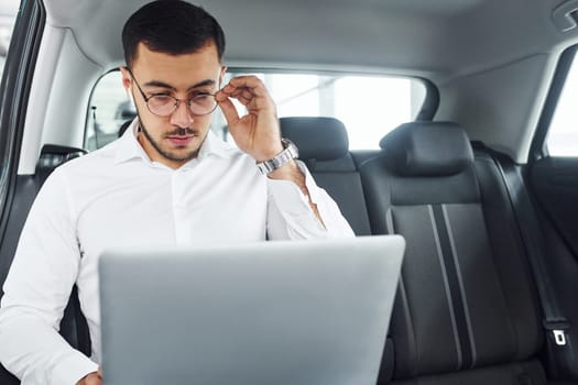 Works by using laptop. Young man in white shirt is sitting inside of a modern new automobile