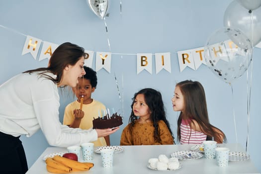 Woman holds cake. Children on celebrating birthday party indoors have fun together